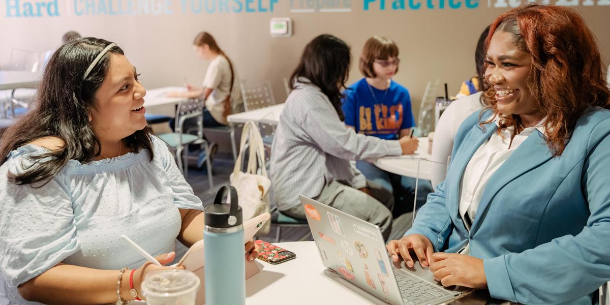 Two students chat and smile while working on a laptop and a tablet